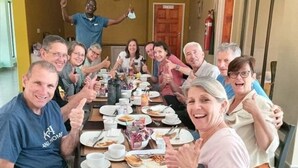 Eine Gruppe von Personen sitzt an einem Tisch mit Tellern voller Essen, ein Feuerlöscher und eine Tür sind im Hintergrund zu sehen.