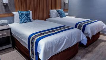 Dos camas con sábanas blancas y almohadas geométricas azules, un cabecero de madera, una mesita de noche y una lámpara de pared.