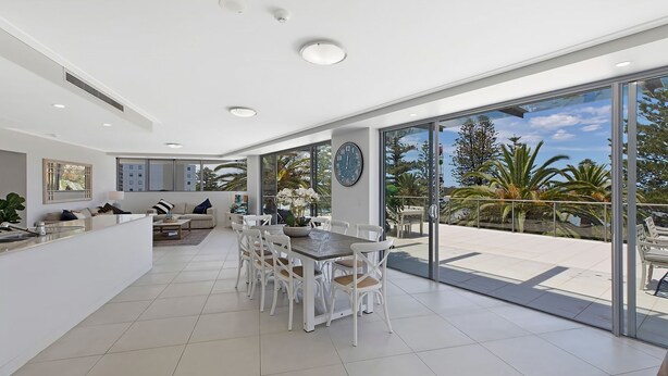 A modern interior with a dining area, kitchen, and large sliding glass doors leading to a balcony with outdoor views. - Coast Luxury Apartments 17 (The Entrance)