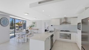 A modern kitchen with a central island, stainless steel appliances, and a dining area with white furniture. - Coast Luxury Apartments 17 (The Entrance)