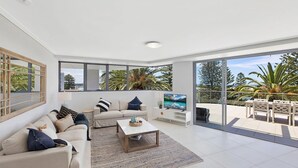 A modern living room with a sofa, coffee table, TV, and a balcony with outdoor furniture. - Coast Luxury Apartments 17 (The Entrance)