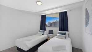 A hotel room with two single beds, a window with curtains, a lamp, and a view of buildings. - Coast Luxury Apartments 17 (The Entrance)