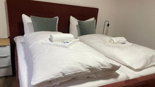 A neatly made bed with white linens and two pillows, a wooden headboard, and a bedside table with a lamp. - Appartio-60m2-6 Pers-netflix (Hofheim in Unterfranken)