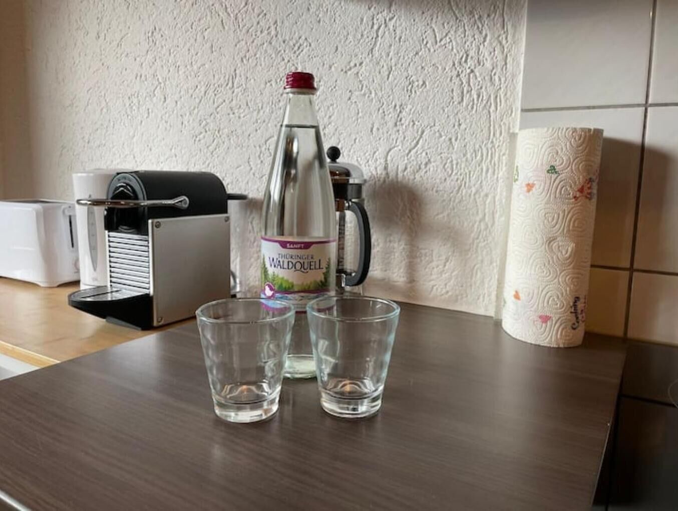 A kitchen counter with a bottle of Thorner Waldquell water, two glasses, a coffee machine, and a paper towel holder.
