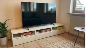 A flat-screen TV on a white entertainment unit with storage drawers, a potted plant, and a view of a building outside a window. - Appartio-60m2-6 Pers-netflix (Hofheim in Unterfranken)