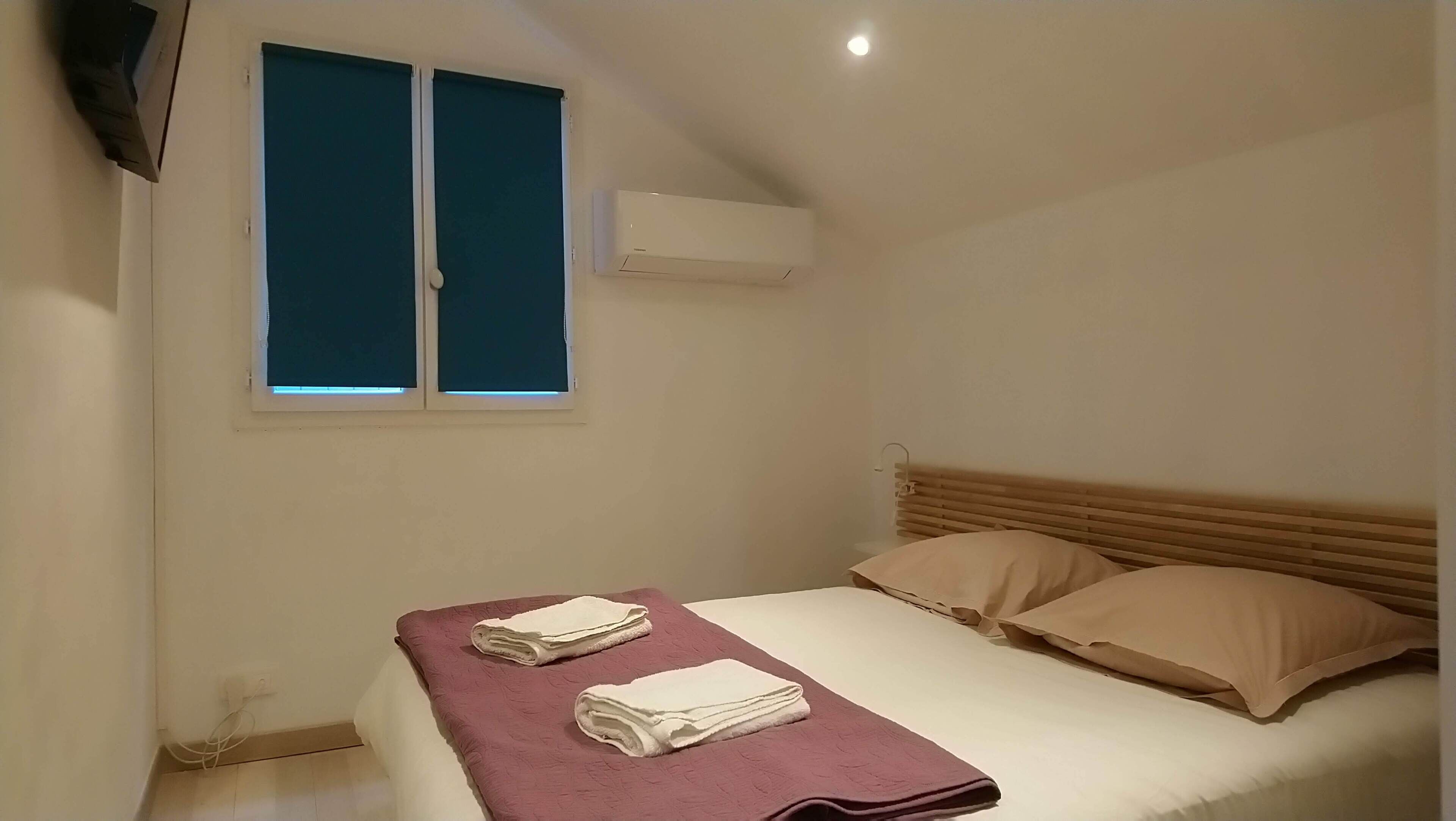 Une chambre à coucher avec un lit, deux oreillers, une fenêtre avec des stores, un appareil de climatisation et une serviette pliée posée sur le lit.