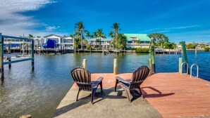 A waterfront dock with two chairs, a clear blue sky, and buildings in the background. - 18116 Cutlass Cozy Pet Friendly Canalfront Home (Fort Myers Beach)
