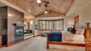 Een ruime slaapkamer met een groot bed, een inbouwplaats voor een open haard, een plafondventilator en uitzicht op het bos door grote ramen.