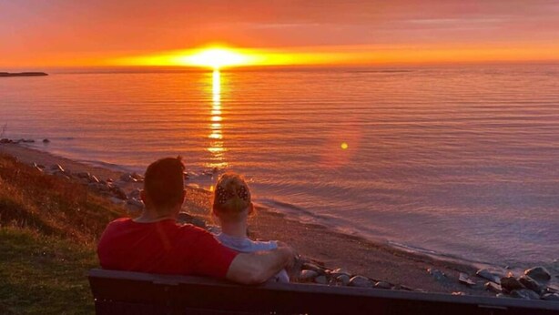 Kaksi ihmistä istuu penkillä ja katselee auringonlaskua, joka heijastuu vesistöstä.