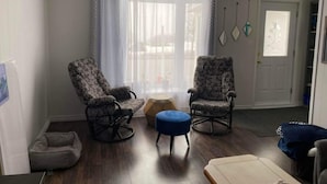 Olohuoneessa on kaksi kuviollinen nojatuolia, pieni jalkarah, koiranpeti ja ovi, josta on näkymä ikkunan kautta.