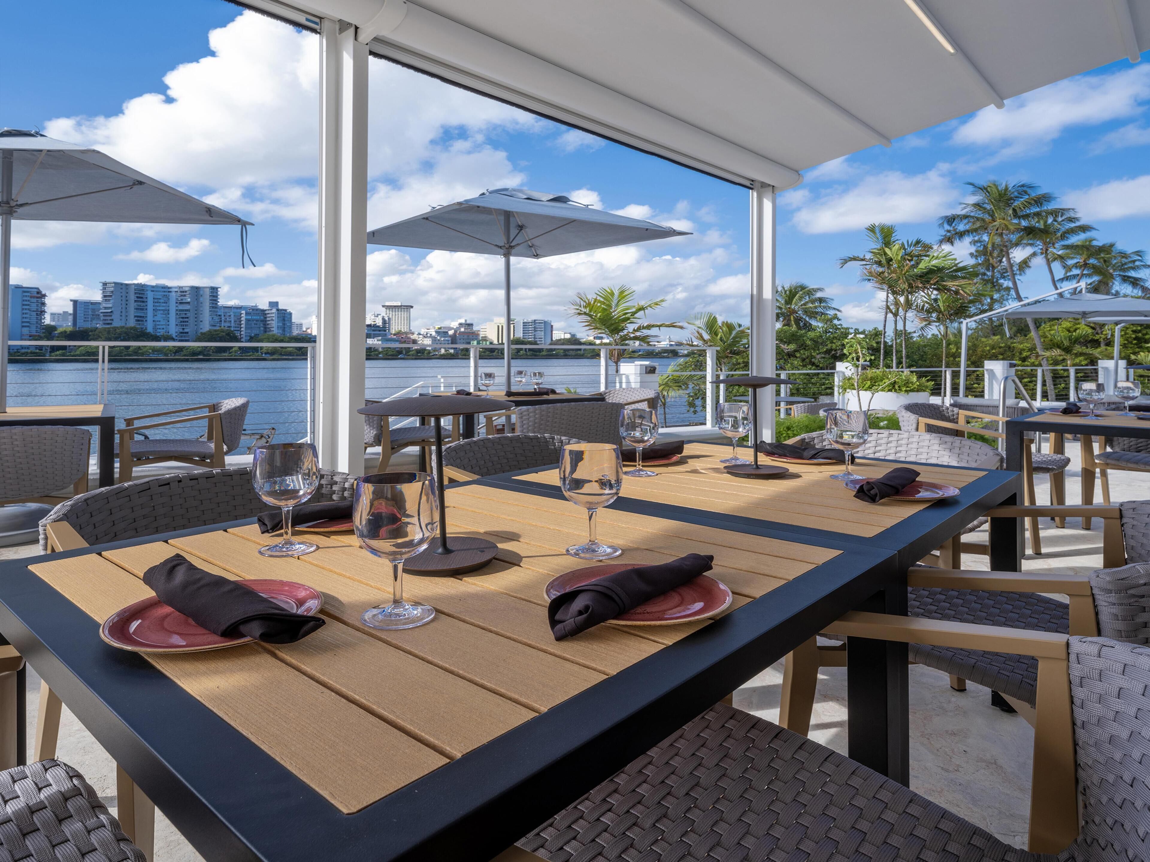 An outdoor dining area with tables and chairs, umbrellas, and a view of the water and city skyline.