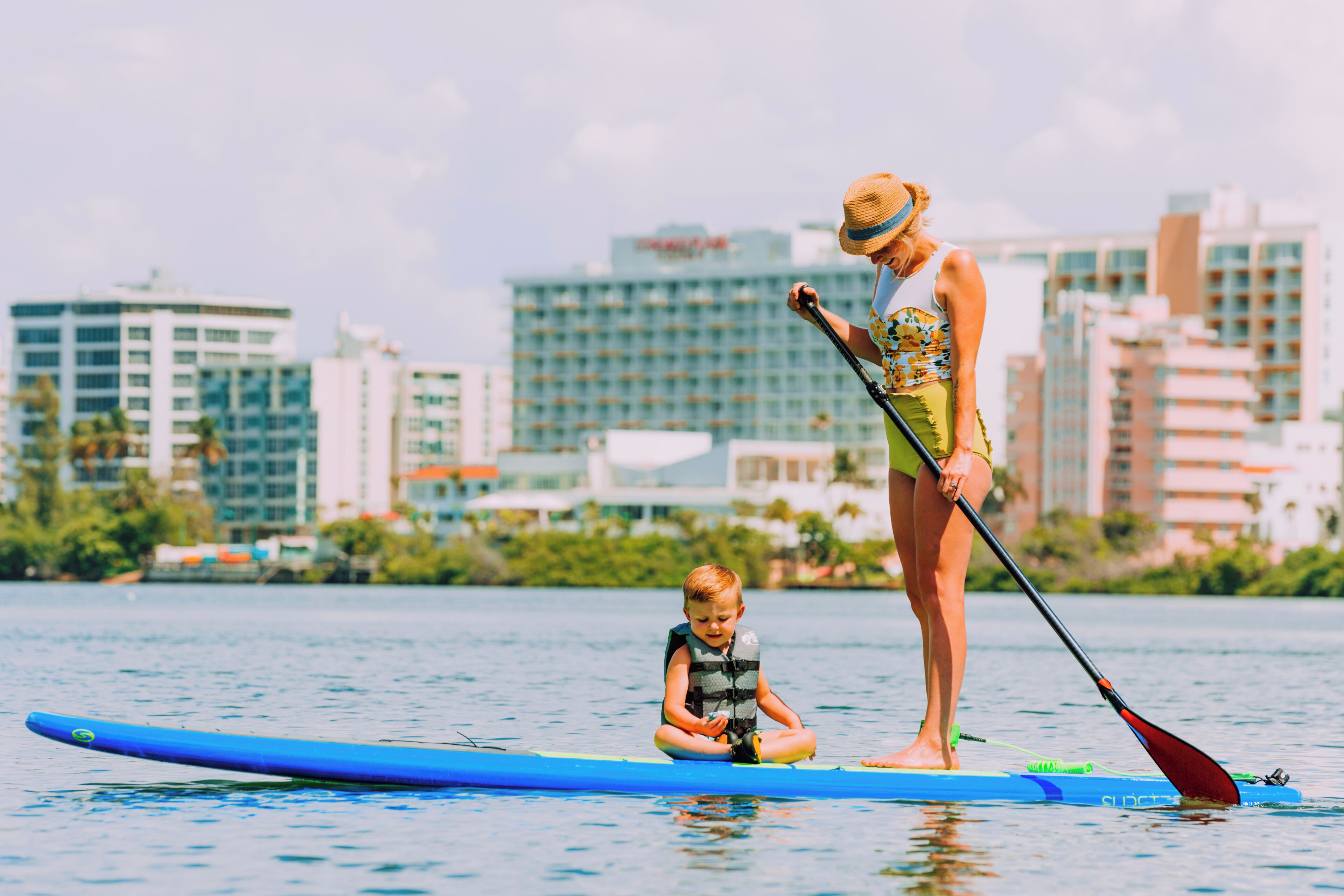 A woman and a child on a paddleboard in a body of water with a cityscape in the background.