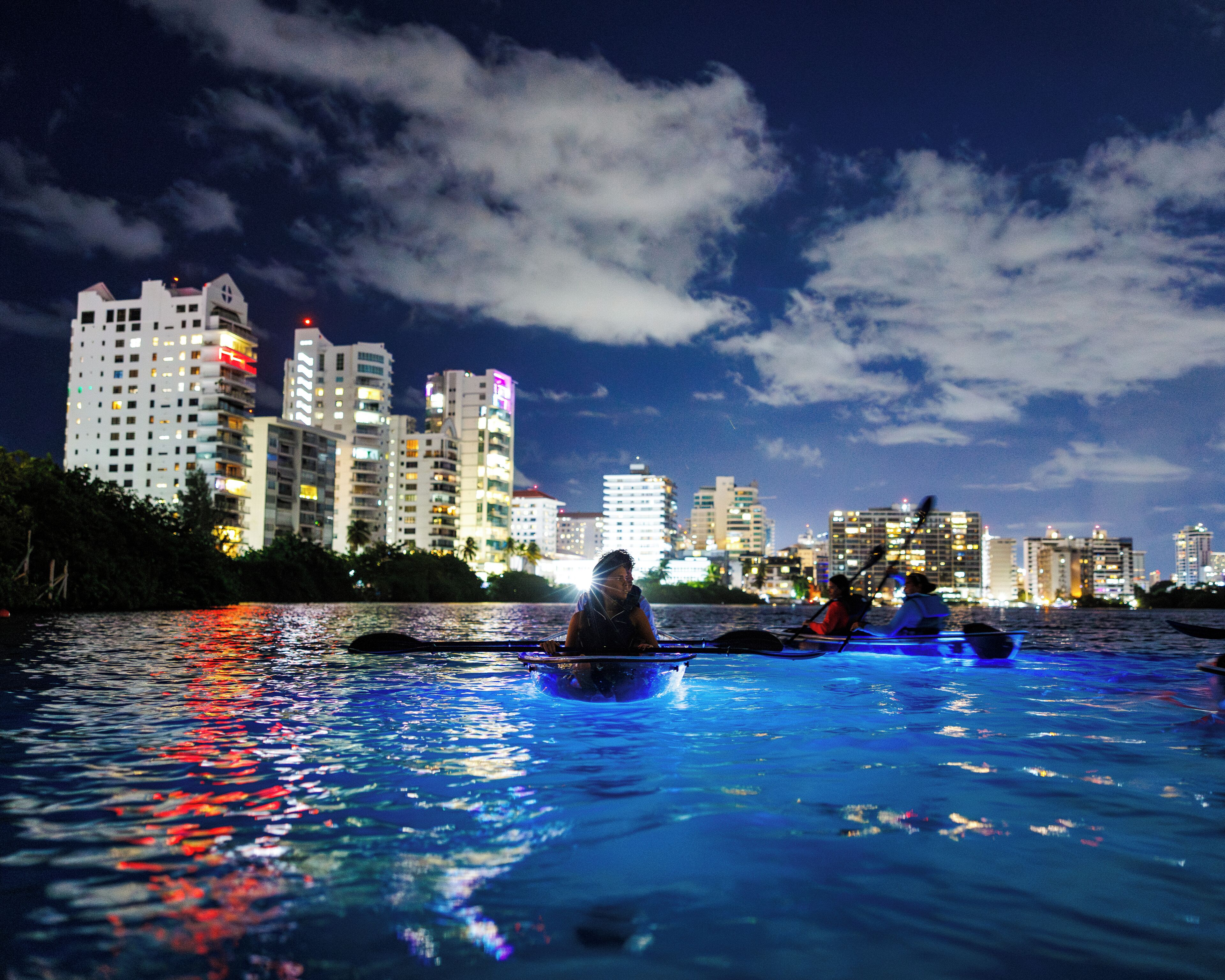 Un paisaje urbano nocturno con edificios al fondo y una masa de agua al frente.
