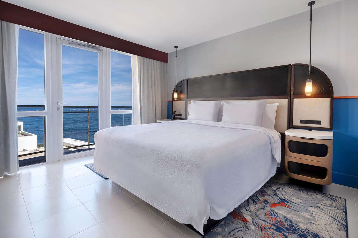 A hotel room with a large bed, a nightstand, a lamp, a view of the sea, and a sliding door leading to an outdoor balcony.