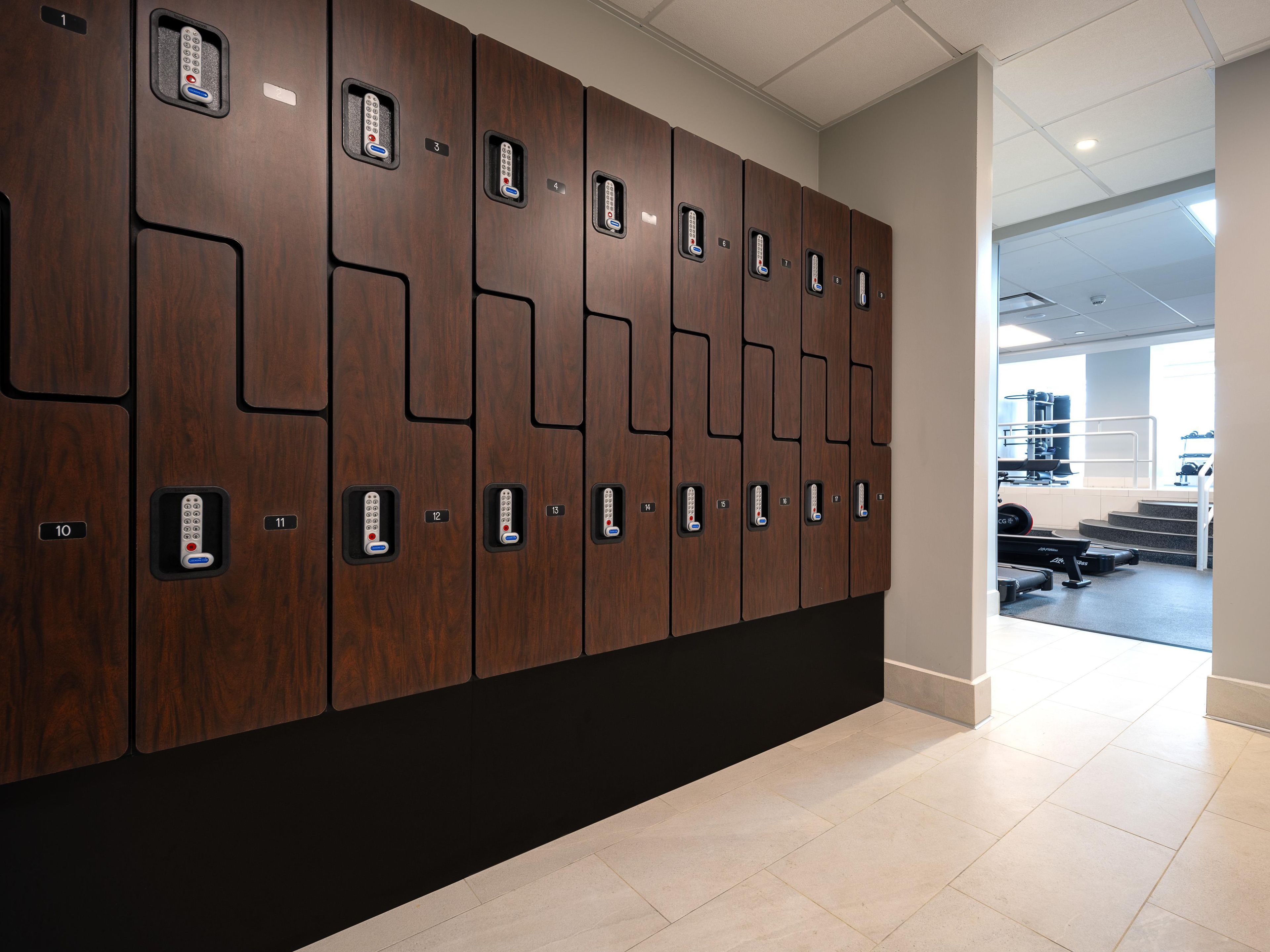 A row of lockers with numbered compartments, a tiled floor, and a glimpse of a gym area in the background.