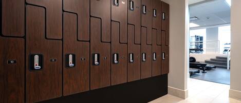 A row of lockers with numbered compartments, a tiled floor, and a glimpse of a gym area in the background.