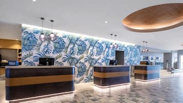 Eine moderne Hotelhalle mit einer Rezeption, einer Wand mit großem tropischen Blattmuster und einem Check-in-Schalter.
