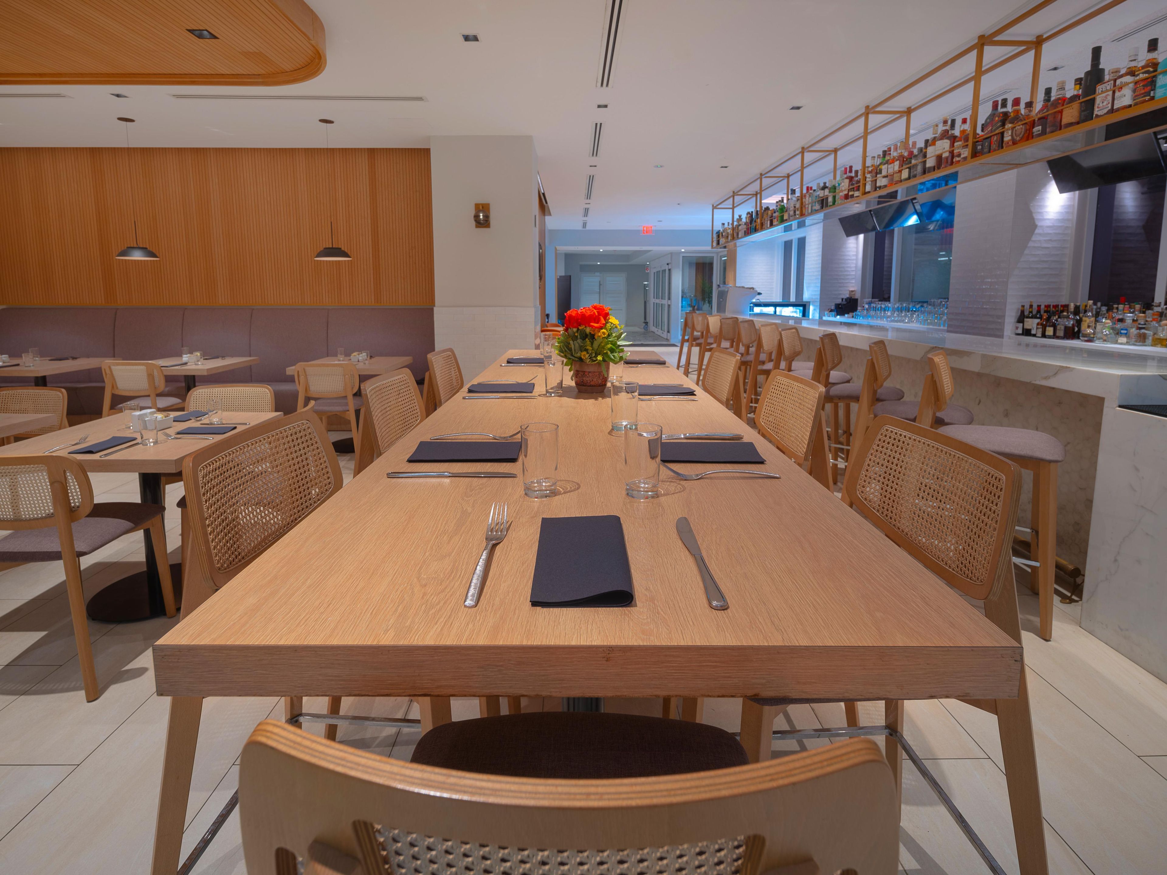 A restaurant with a long wooden table set for dining, surrounded by chairs, and a bar area with shelves of bottles.