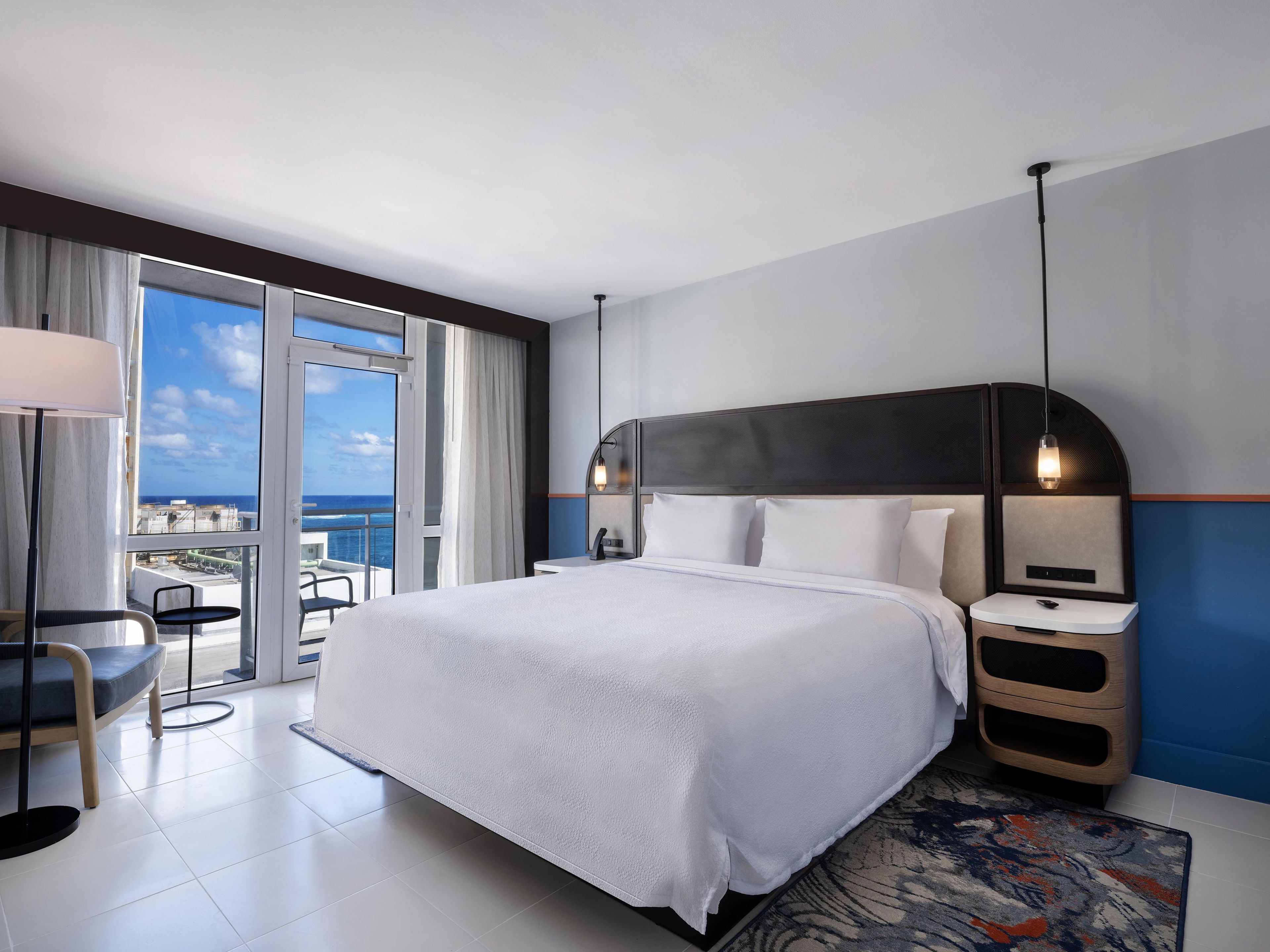 A modern hotel room with a large bed, bedside tables, a desk, and a view of the ocean.