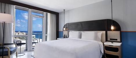 A modern hotel room with a large bed, bedside tables, a desk, and a view of the ocean.