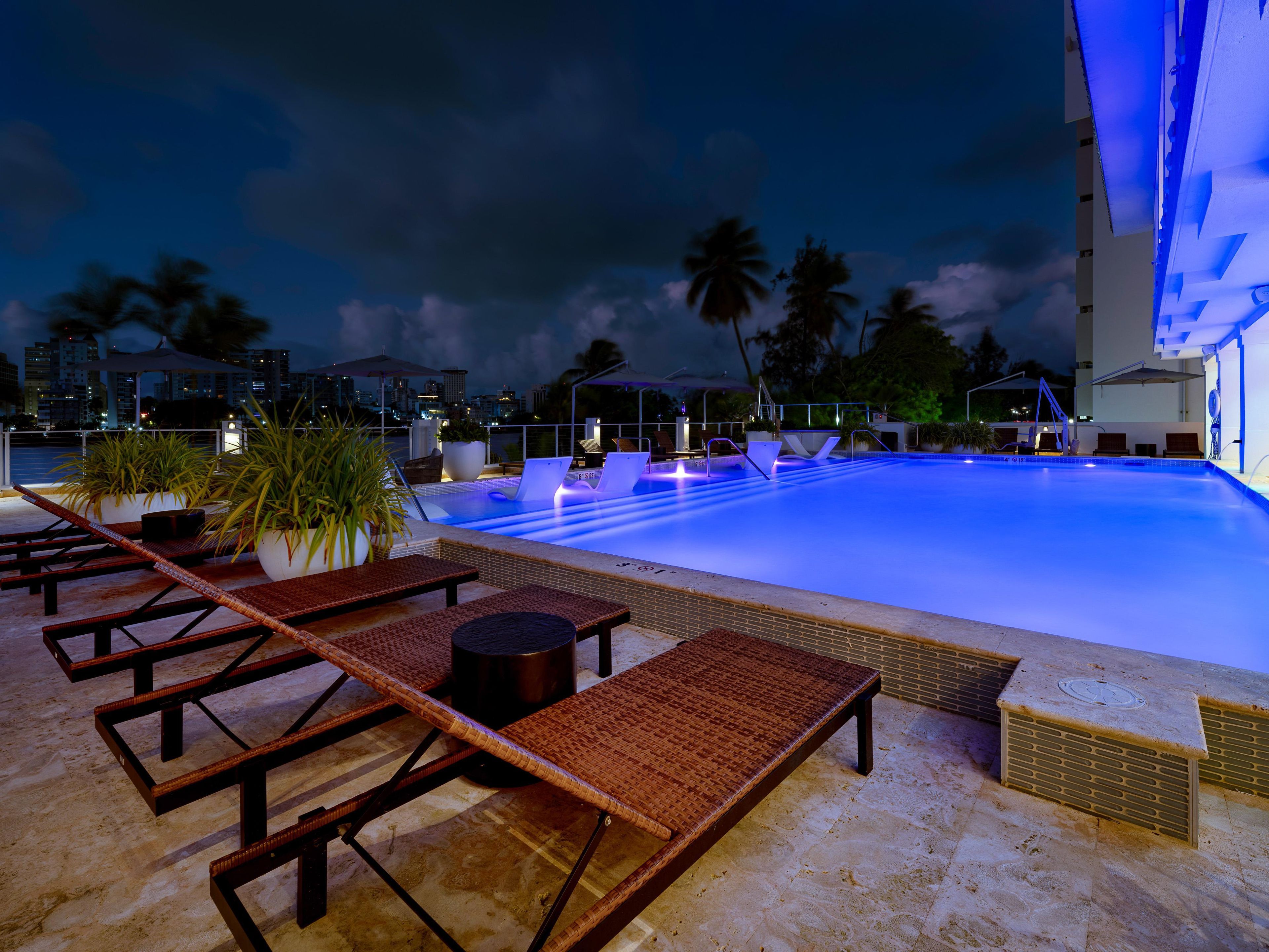 A swimming pool with lounge chairs and potted plants, surrounded by palm trees and a city skyline at dusk.
