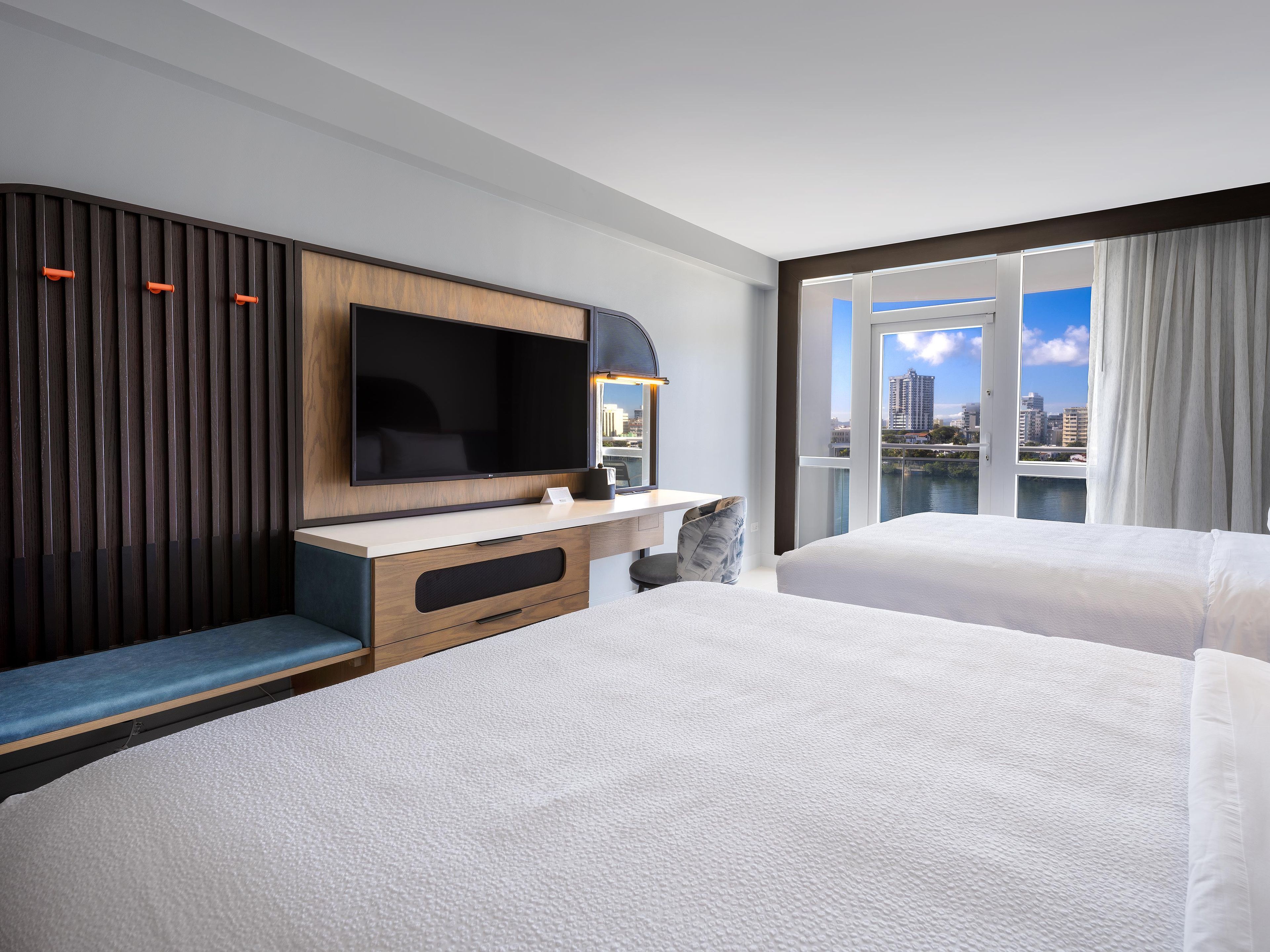 Habitación de hotel con una televisión de pantalla plana grande, un escritorio con una silla, un sofá y un balcón con vista a la ciudad.