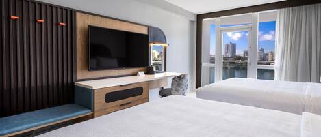 A hotel room with a large flat-screen TV, a desk with a chair, a sofa, and a balcony with a city view.