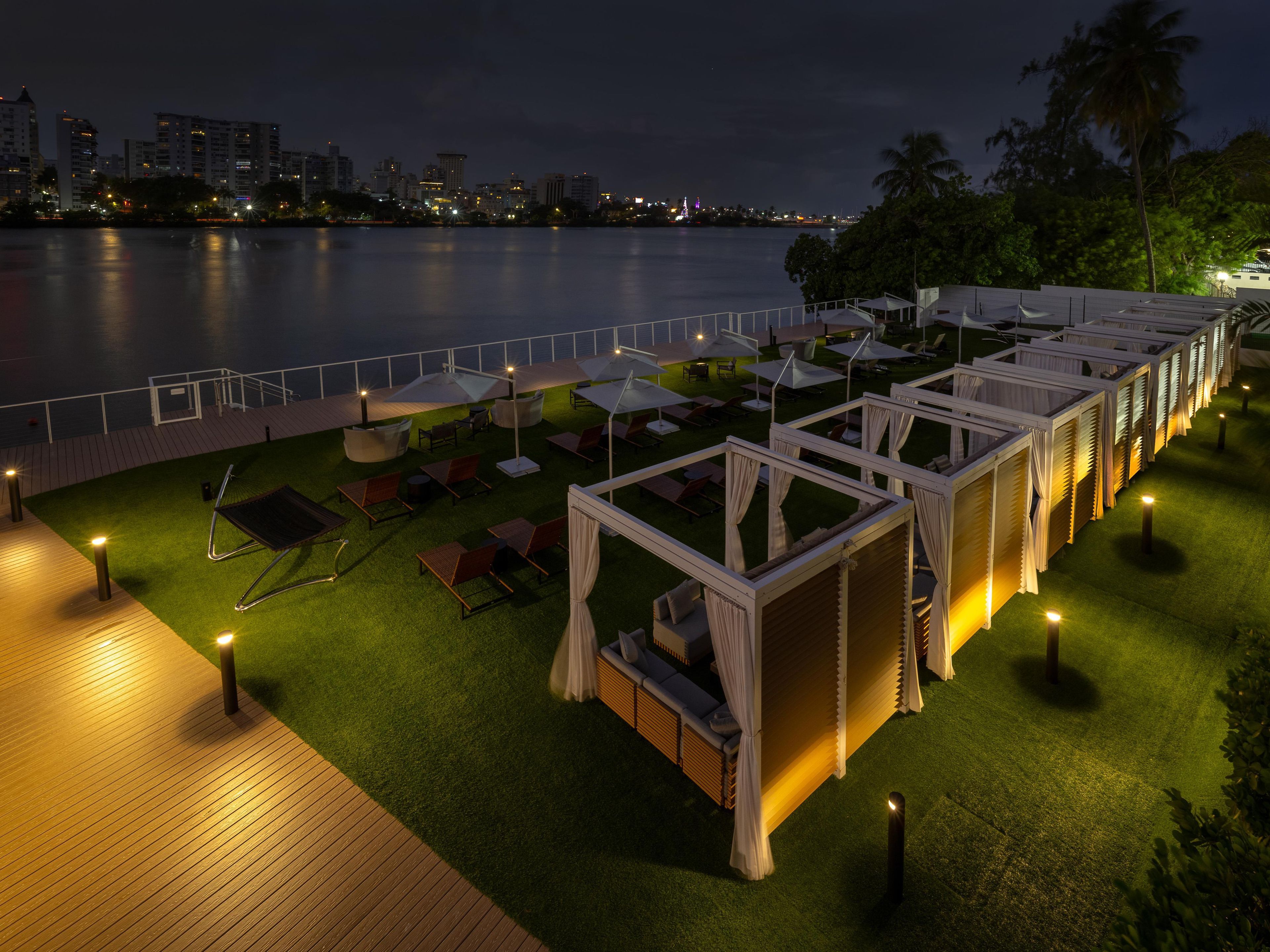 An outdoor seating area with umbrellas and lounge chairs, a wooden deck, and a view of the water and city skyline at night.