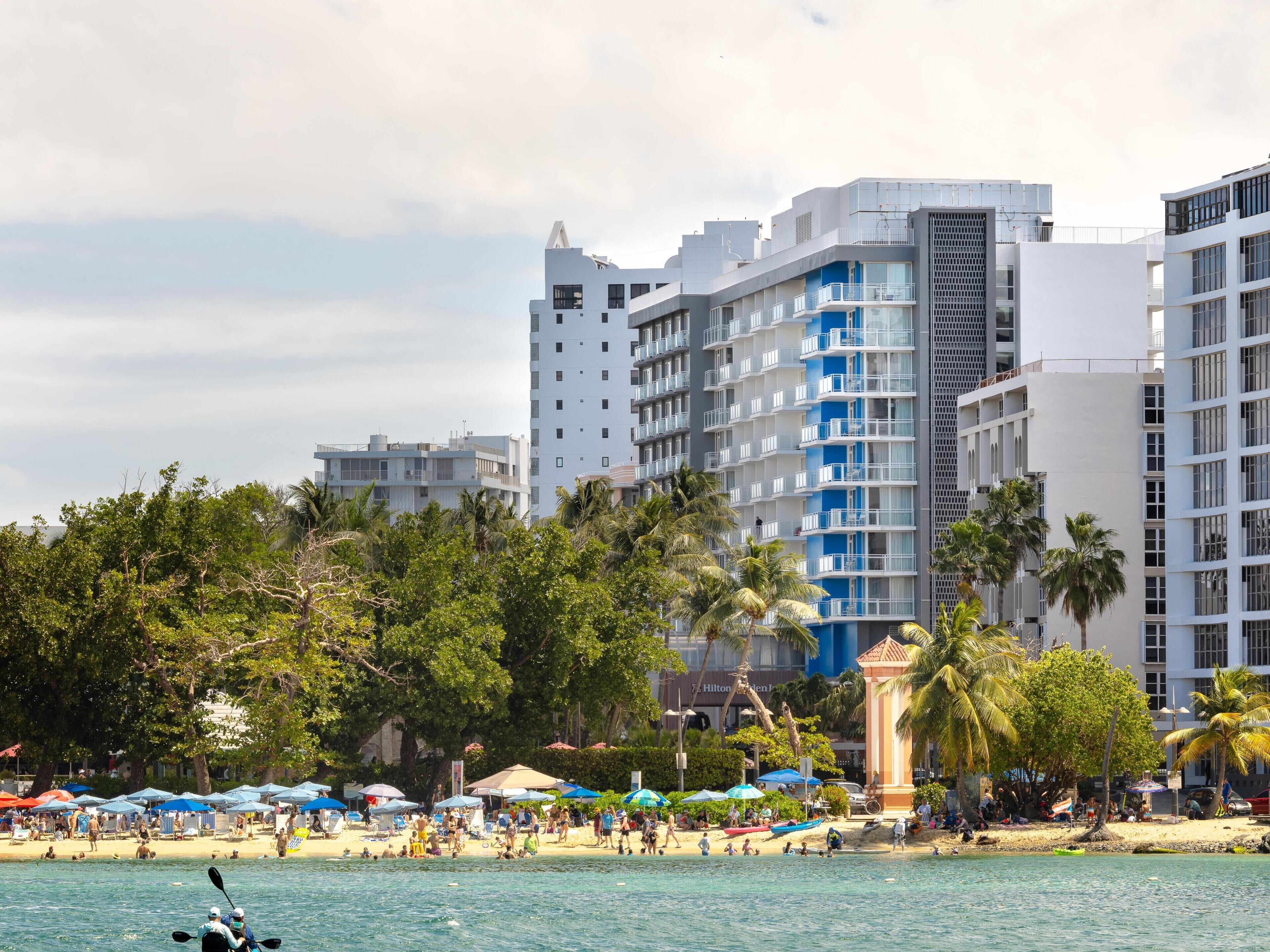 A beachfront hotel with multiple floors, a beach in the foreground, and palm trees.