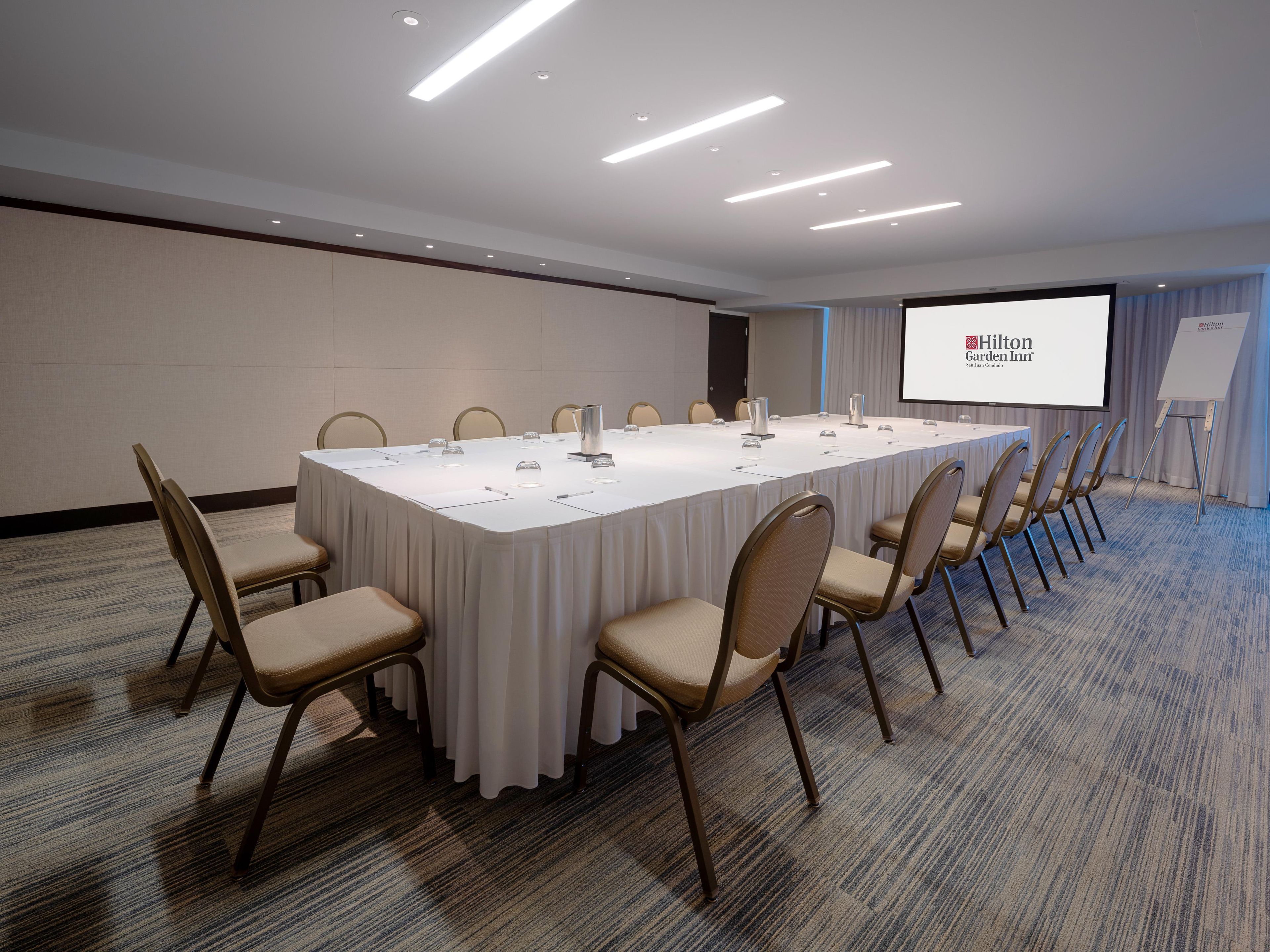 Un salón de conferencias con una larga mesa, sillas, una pantalla de proyector y una pizarra.