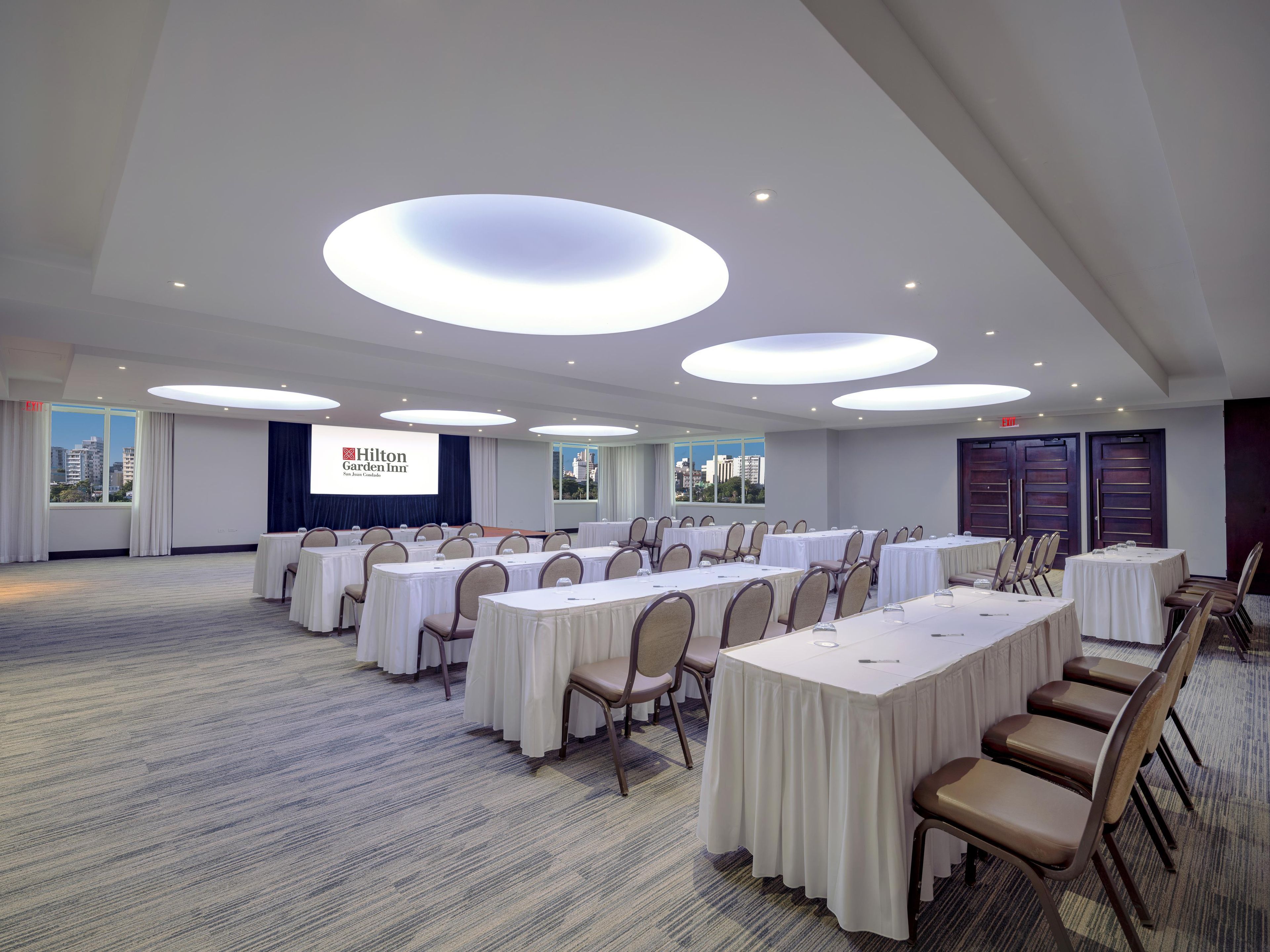 A conference room with multiple tables and chairs, a large screen displaying the Hilton Garden Inn logo, and a city view through the windows.