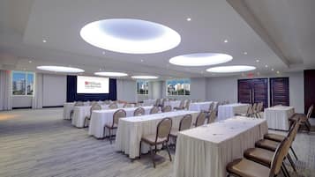 A conference room with multiple tables and chairs, a large screen displaying the Hilton Garden Inn logo, and a city view through the windows.