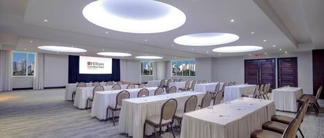 A conference room with multiple tables and chairs, a large screen displaying the Hilton Garden Inn logo, and a city view through the windows.