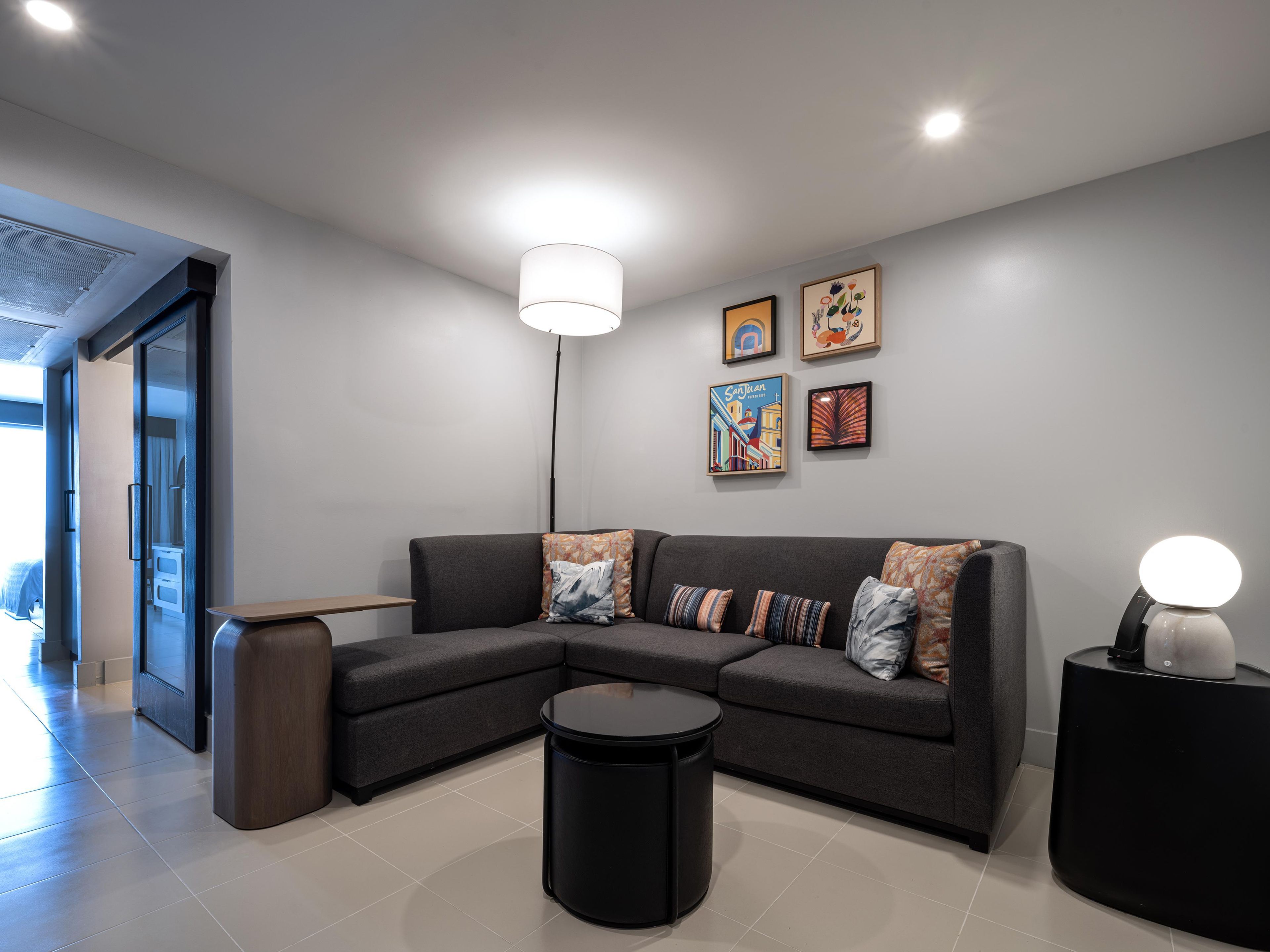 Een moderne woonkamer met een hoekbank, een ronde salontafel en een bijzettafeltje met een lamp. Er hangen lijstwerken aan de muur en een deur die leidt naar een andere kamer.