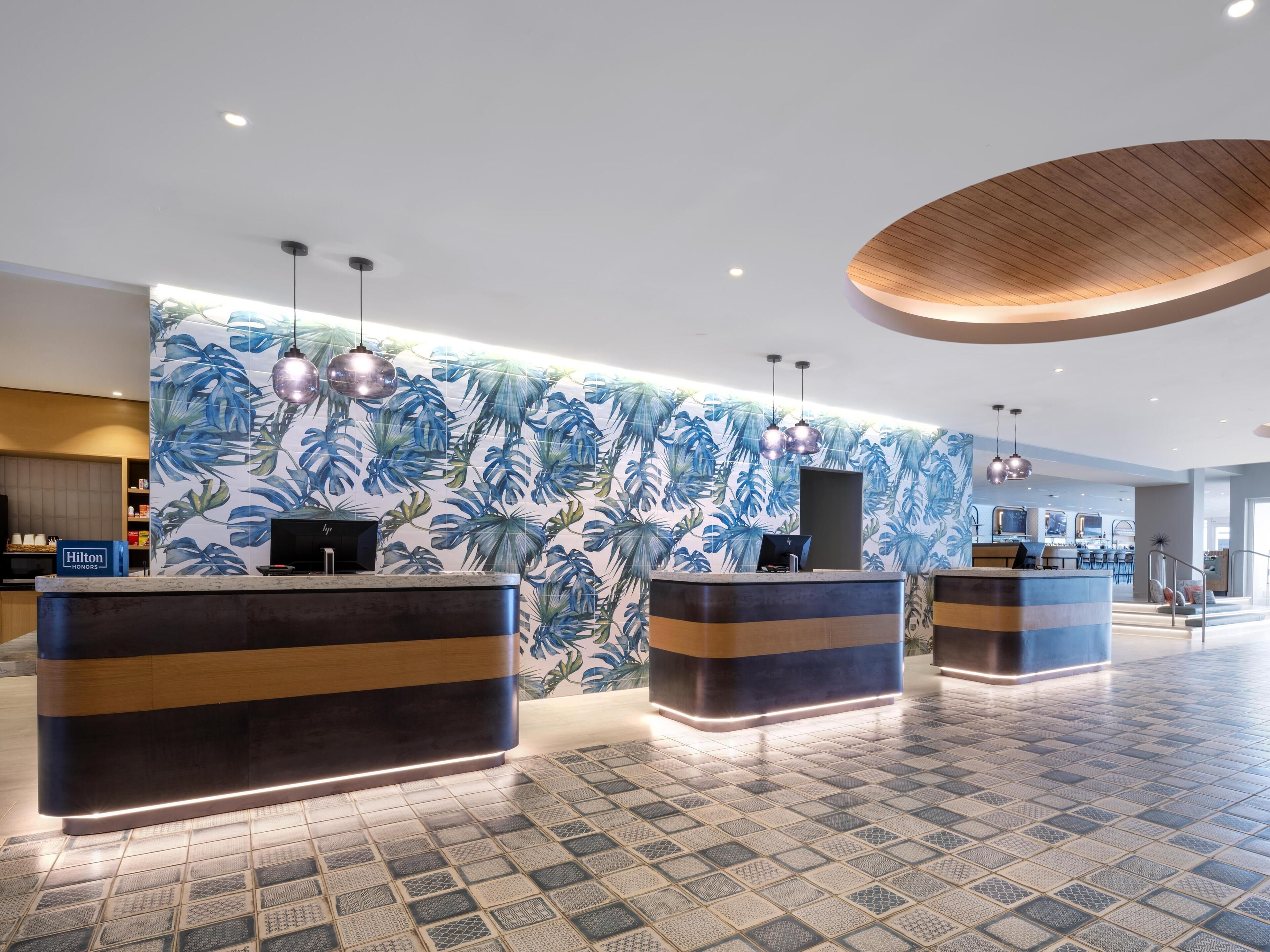 En modern hotellobby med en receptionsdisk, en stor vägg dekorerad med ett tropiskt bladmotiv och en incheckningsdisk.