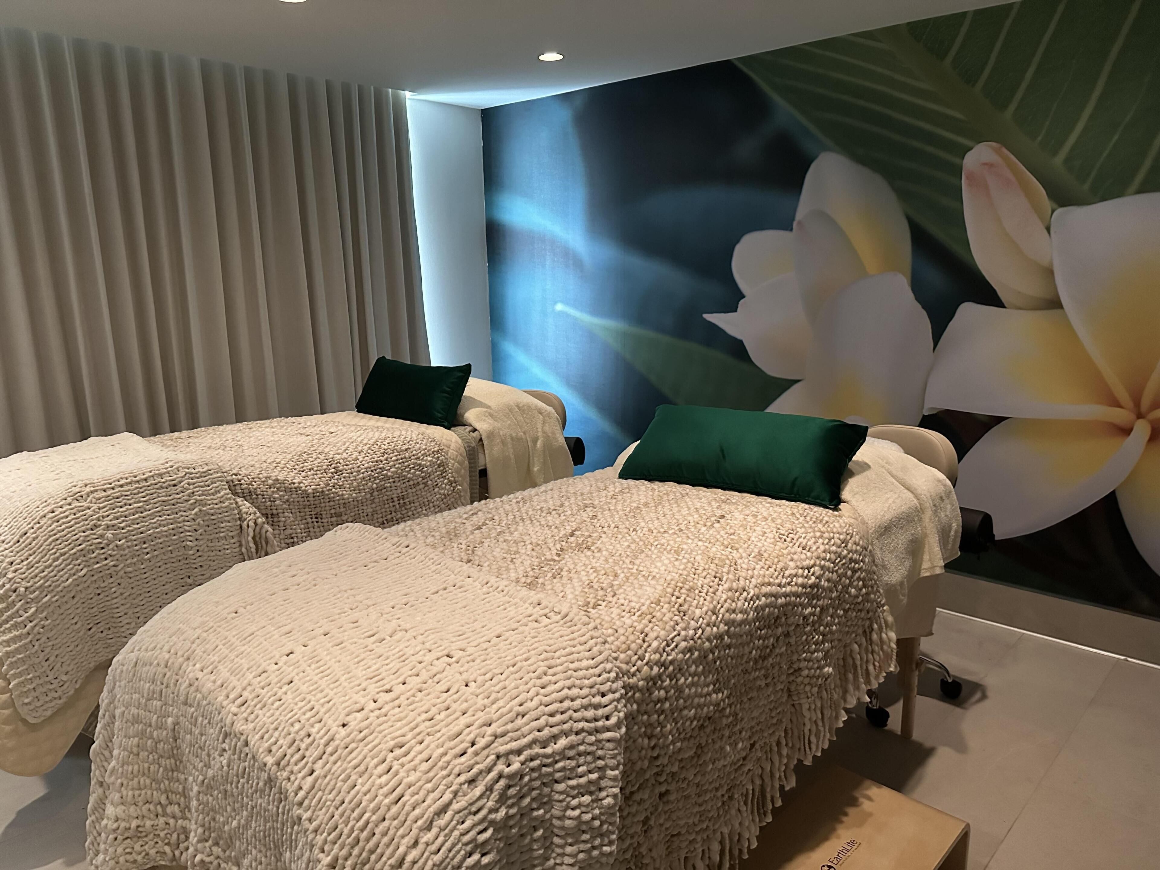 A spa room with two massage tables, beige textured blankets, green pillows, and a large floral mural on the wall.