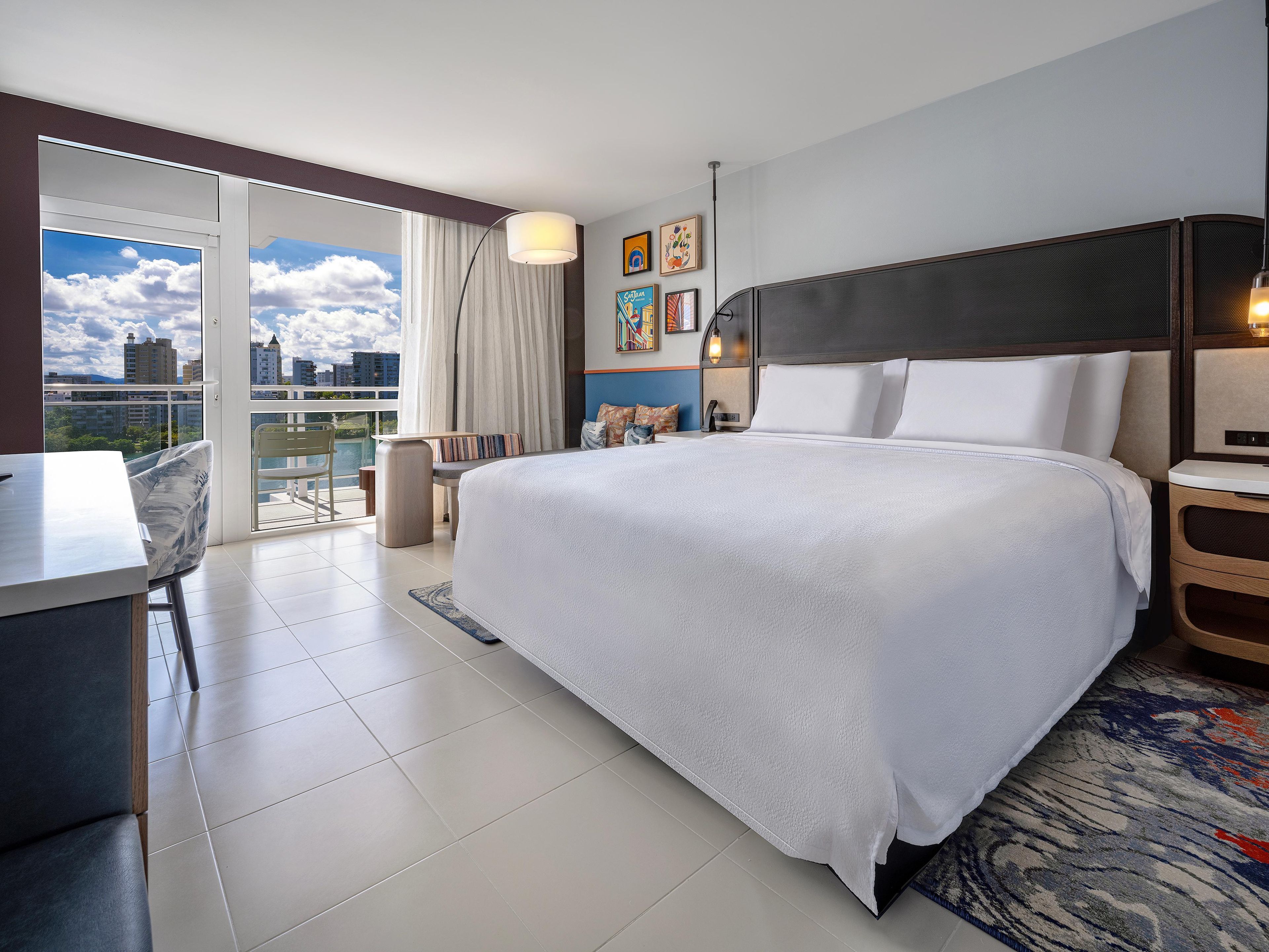 Quarto de hotel com uma cama grande, uma escrivaninha, uma cadeira, varanda com vista para a cidade e obras de arte nas paredes.