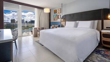 Quarto de hotel com uma cama grande, uma escrivaninha, uma cadeira, varanda com vista para a cidade e obras de arte nas paredes.