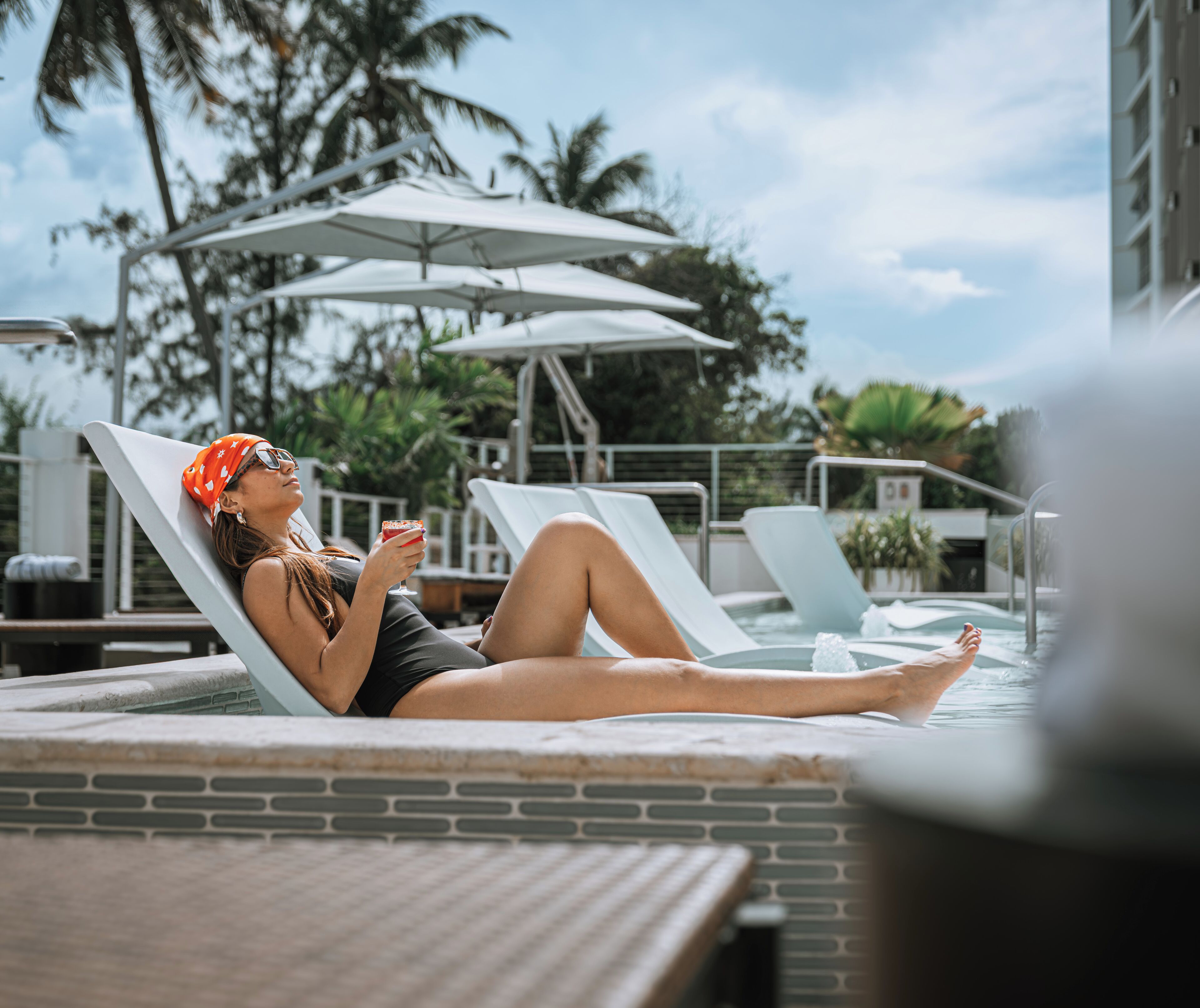 Un espace piscine équipé de chaises longues, de parasols, et où une personne se détend en sirotant une boisson.