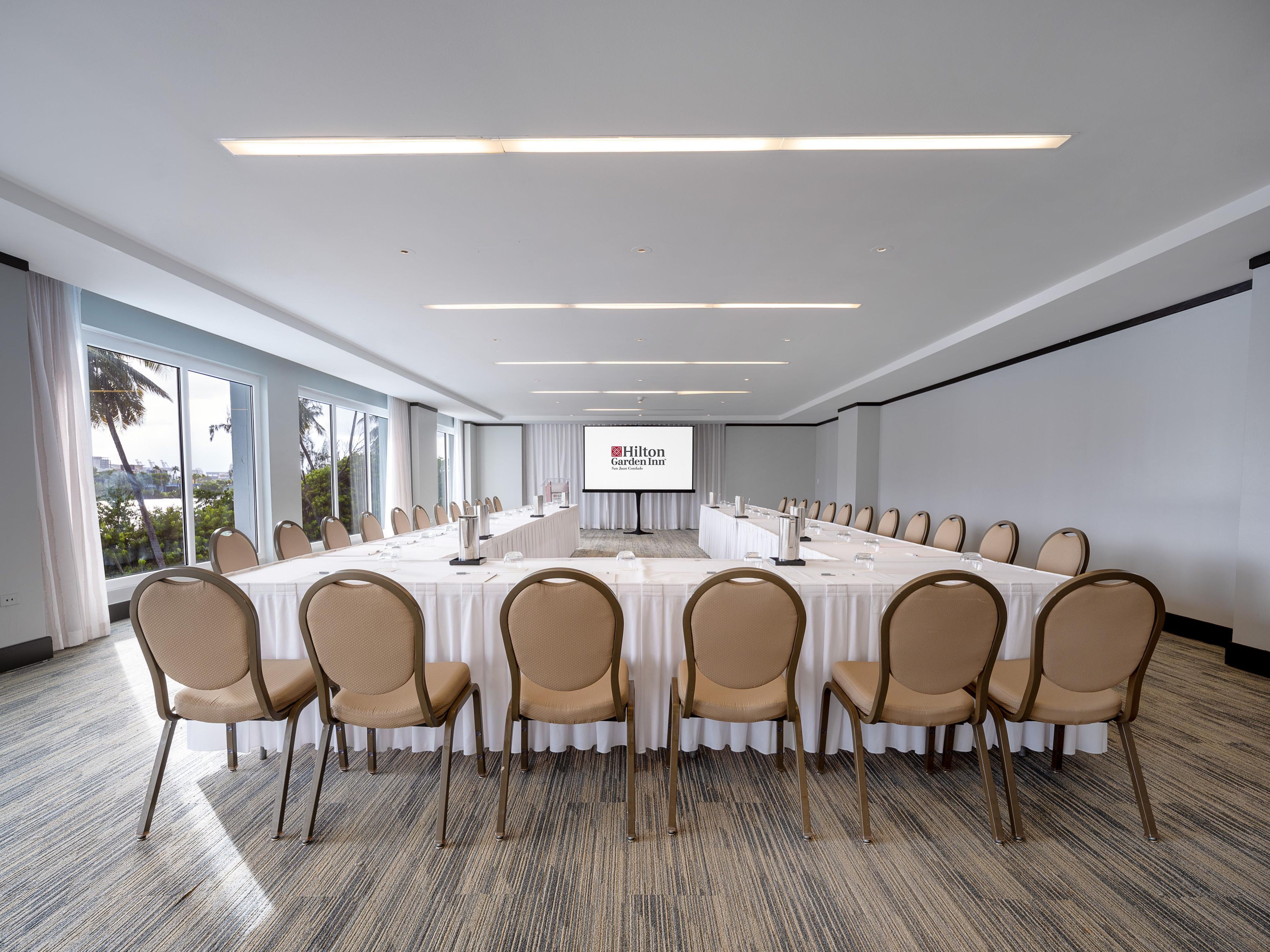 A conference room with a long table, chairs, a projector screen, and a view of palm trees through large windows.