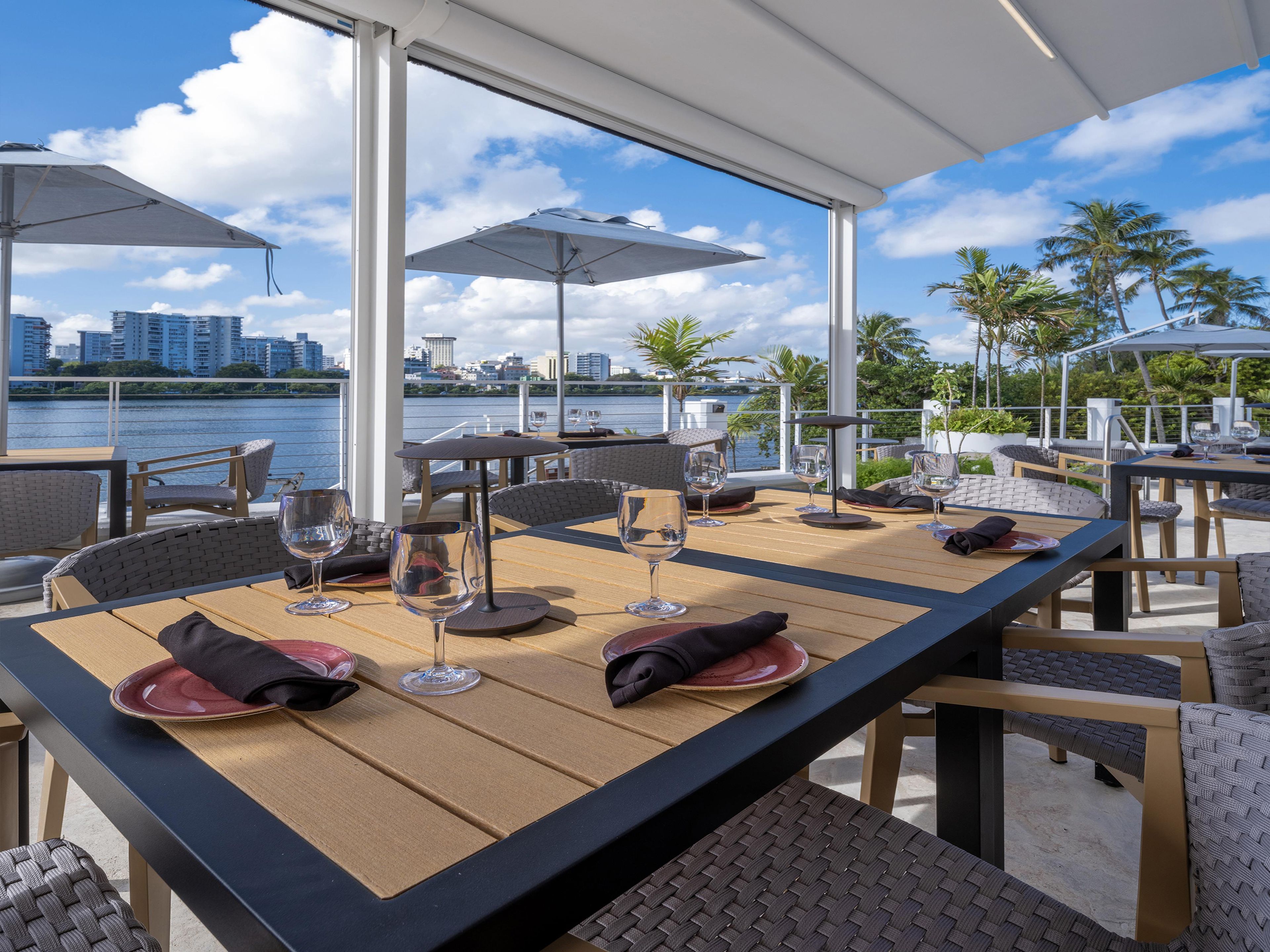 An outdoor dining area with tables and chairs, overlooking a body of water and a city skyline.