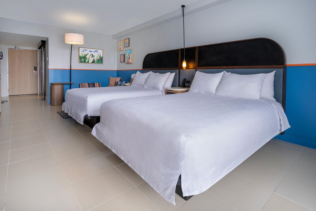 A hotel room with two beds, a nightstand, a lamp, and artwork on the walls.