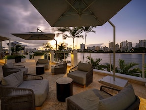 An outdoor seating area with wicker furniture, umbrellas, and a view of the city skyline at dusk. - Hilton Garden Inn San Juan Condado (San Juan)