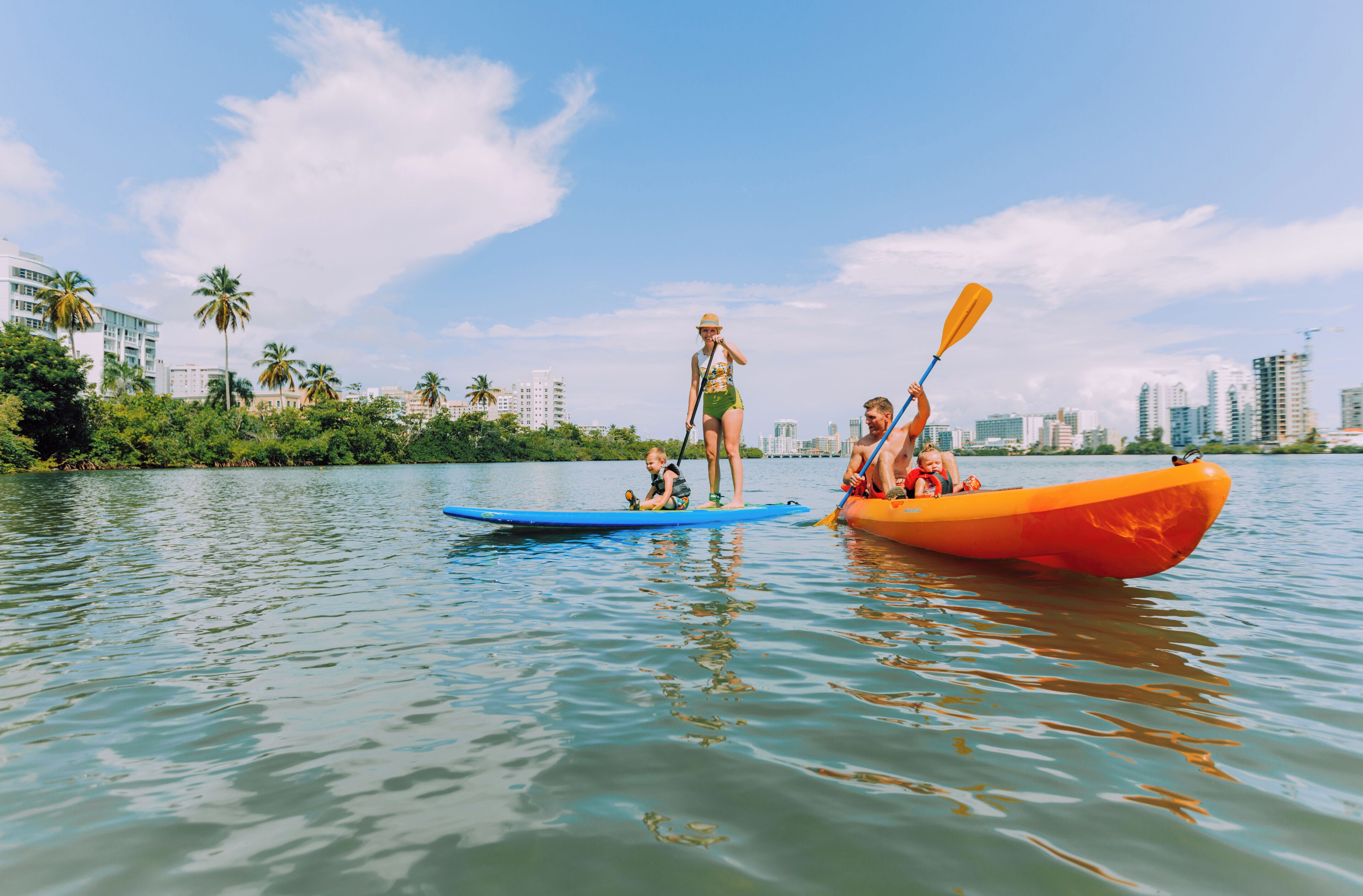 Un río tranquilo con kayaks, una persona de pie en una tabla de remo y un paisaje urbano al fondo.