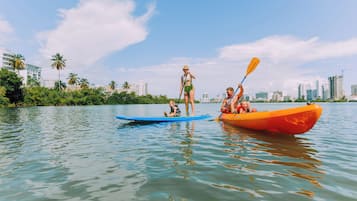 Un río tranquilo con kayaks, una persona de pie en una tabla de remo y un paisaje urbano al fondo.