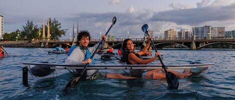 Two people in a kayak on a body of water with a bridge and buildings in the background.