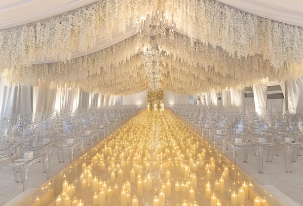 Um longo corredor iluminado por velas, com cadeiras dispostas de ambos os lados, conduzindo a uma área central com um candelabro e tecidos pendurados.