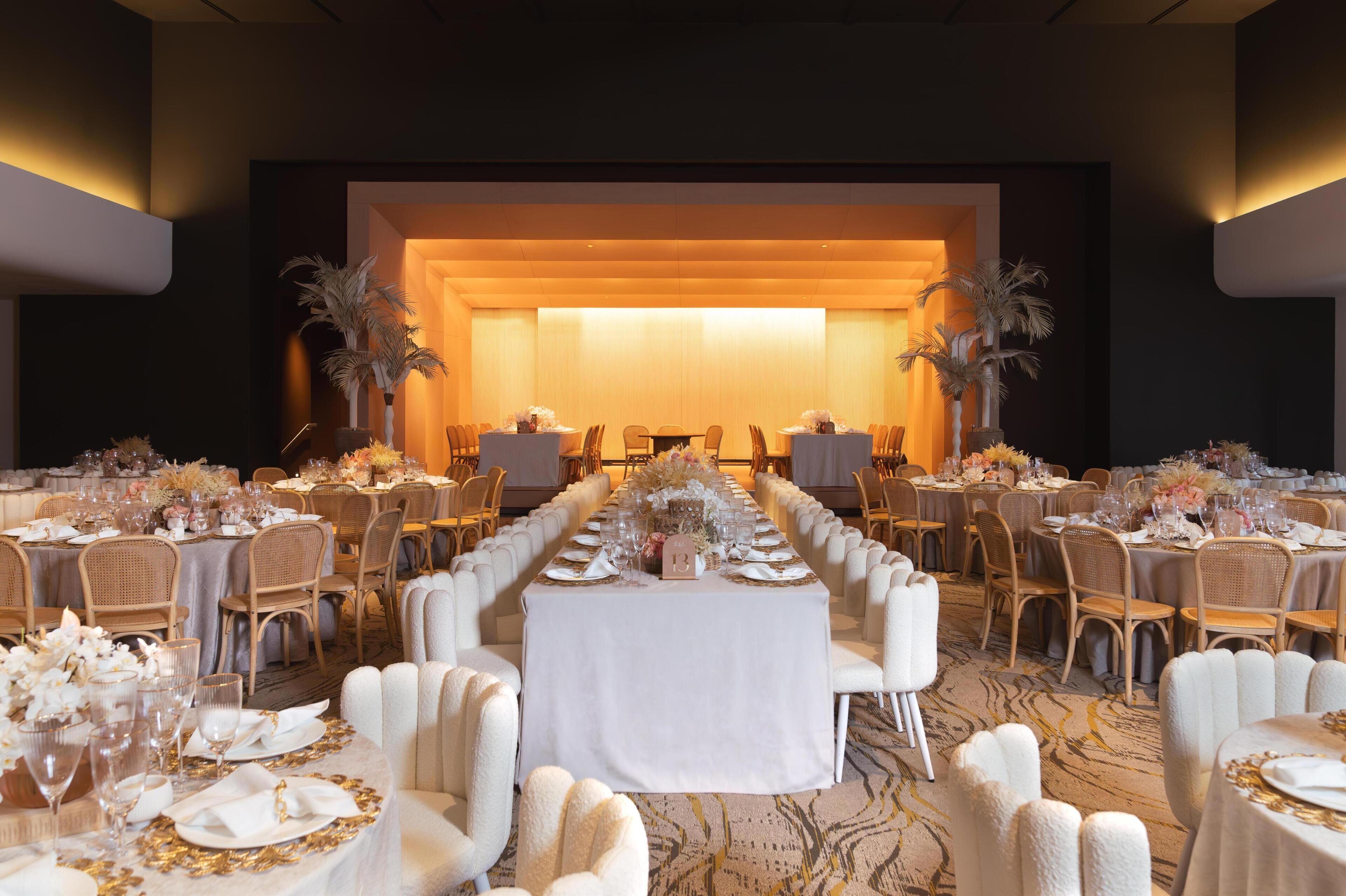 A banquet hall with multiple round tables, each set with white tablecloths, gold accents, and floral centerpieces.