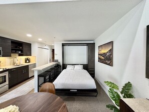 A compact hotel room with a bed, a small kitchenette, a desk, and a view of a mountain.