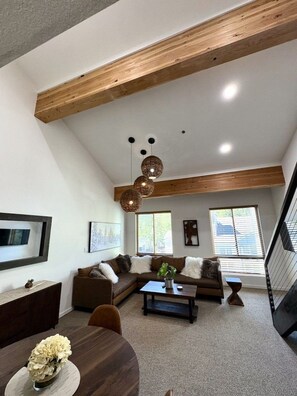 A loft-style living space with exposed wooden beams, a sectional sofa, a dining table, and a large window providing natural light.